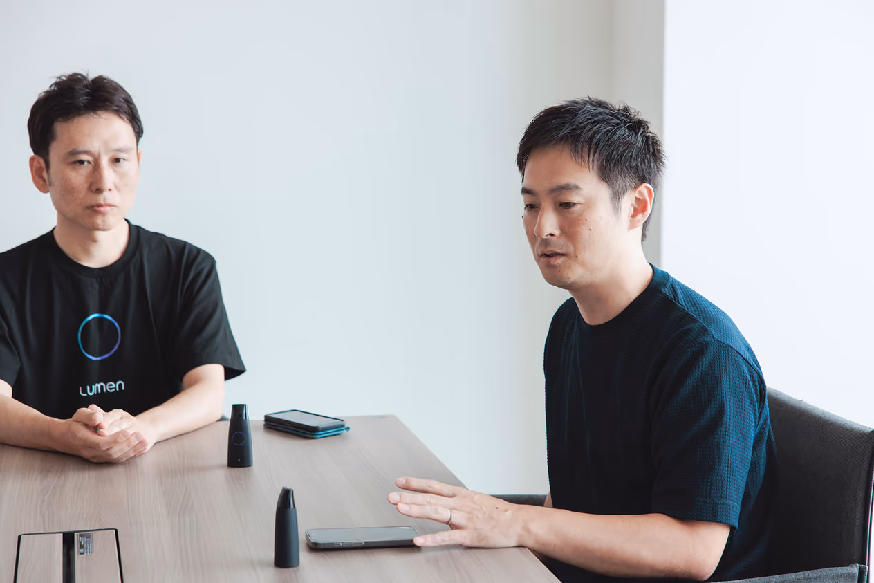 Sho Tsugawa explaining Lumen on a desk, with Yuji Kaneko listening